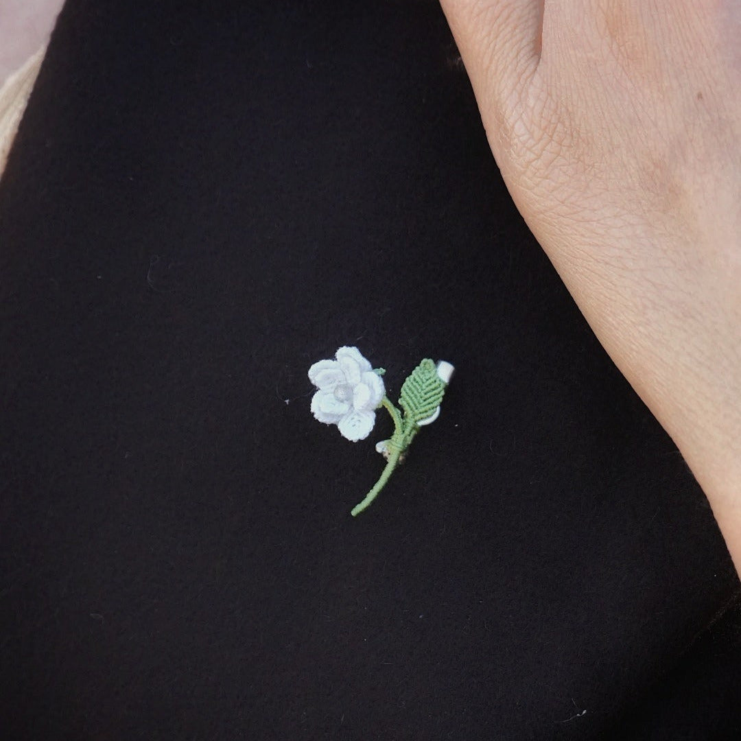 Mini broche fleur blanche en micro macramé avec cœur en labradorite bleu-vert, épinglée sur manteau en laine marron foncé, bijou elfique et bohème fait main.