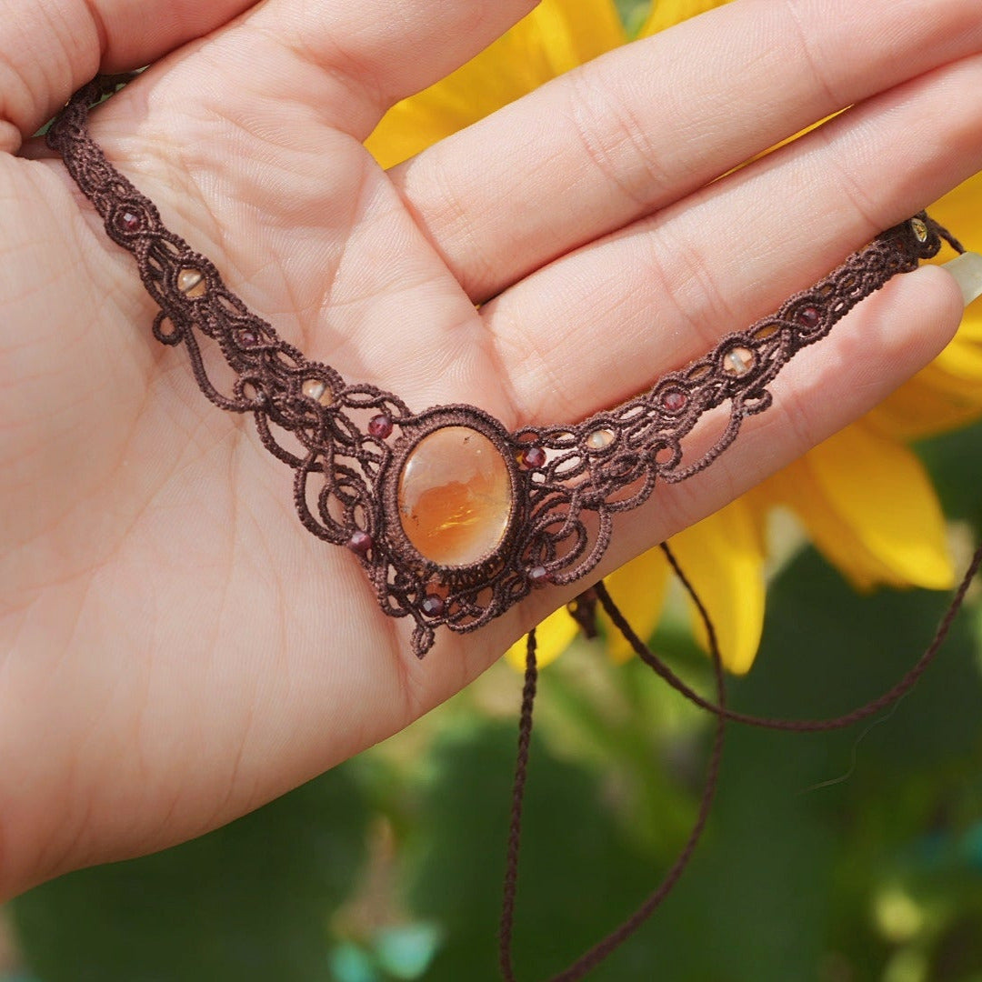 Collier ras-de-cou elfiques en micro macramé avec pierre naturelle. Bijoux tissé en fil de coton de petit diamètre. Mini Grenats rouge rosé facettés autour d'une citrine jaune ambré ovale, fil de coton marron foncé. Motifs tissés en entrelacs celtiques et arabesques elfiques Forme V. Bijoux ambiance bohème hippie aux couleurs chaudes. Bijoux posé à la verticale dans une mains à la peau clair avec fond tournesol jaune et nature. Bijoux vue de face