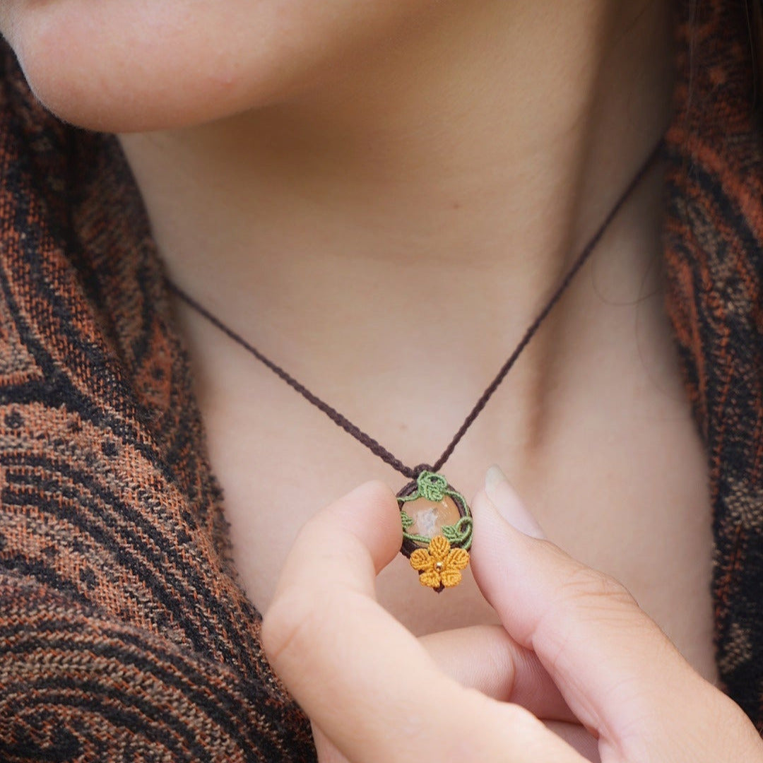 Collier avec pendentif en micro macramé avec motifs floraux. Bijou féminin composé d'un citrine jaune pâle translucide ovale sertie dans une coque marron foncé tissé avec un fil de 0.5 mm sur lequel est fixé une liane verte entrelacée avec mini feuilles et une fleur à 5 pétales jaune orangé avec un cœur doré de 2 mm.
Le bijou est porté autour d'un cou de femme à la peau claire et d'un châle orange et noir. le bijou est tenu entre pouce et index vu de face