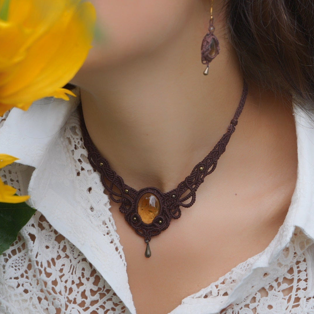 Collier large en micro macramé de couleur marron foncé, jaune ambré et bronze. Motif tissé en micro macramé avec ambiance entrelacs celtiques et arabesques elfiques. Présence d'une pierre centrale ovale de grande taille jaune translucide, gouttes bronze, forme V avec mini perles ronde bronze parsemées. Photo ambiance bohème hippie avec une touche d'élégance. Bijou porté sur une peau claire avec tournesol jaune en premier plan et chemise blanche en dentelle.