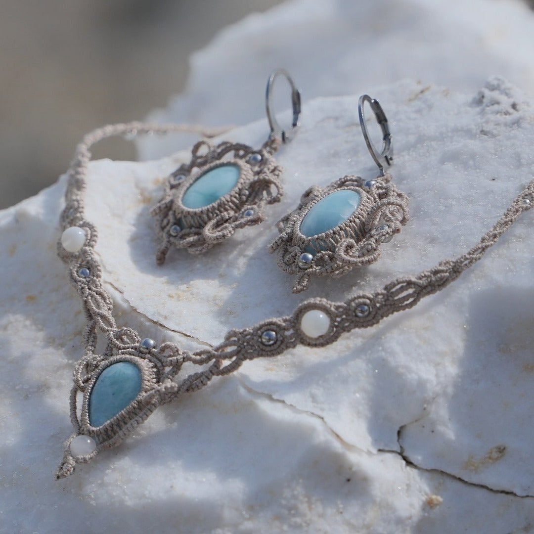 Parure Trésors d'Ecume composée d'une paire de boucles d'oreilles pendentes avec collier ras de cou assorti. Larimar bleu ciel ovales pour les boucles et en poire pointe en bas pour le collier. 3 Perles en nacre blanche pour le collier. Perles acier inoxydable argenté pour les deux pour le côté luxe et précieux. Dormeuses en acier inoxydable pour un port assuré. Fil de Coton couleur lin naturel (beige gris) de 0, 5 mm tissé en micro-macramé en relief version celtique. Bijoux posés sur une pierre blanche