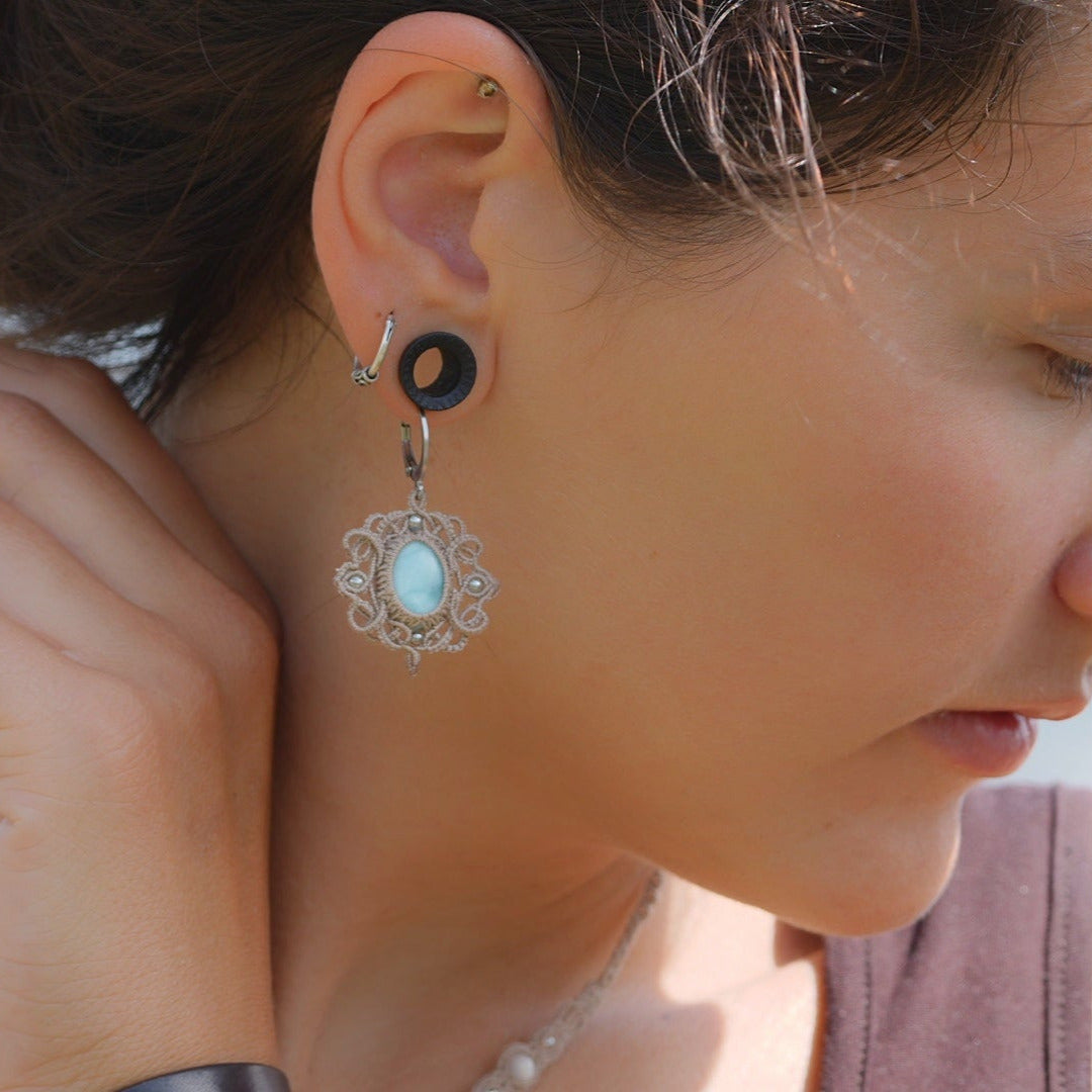 Parure Trésors d'Ecume composée d'une paire de boucles d'oreilles pendentes avec collier ras de cou assorti. Larimar bleu ciel ovales pour les boucles et en poire pointe en bas pour le collier. 3 Perles en nacre blanche pour le collier. Perles acier inoxydable argenté pour les deux pour le côté luxe et précieux. Dormeuses en acier inoxydable pour un port assuré. Fil de Coton couleur lin naturel (beige gris) de 0, 5 mm tissé en micro-macramé en relief version celtique. Bijoux porté femme brune peau caramel