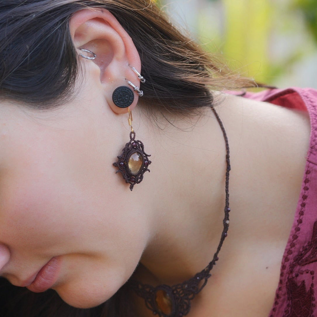 Boucles d'oreilles elfiques en micro macramé avec pierre naturelle. Bijoux tissé en fil de coton de petit diamètre. 4 Mini Grenats rouge rosé facettés autour d'une citrine jaune ambré, fil de coton marron foncé. Motifs tissés en entrelacs celtiques et arabesques elfiques. Crochet doré. Bijou ambiance bohème hippie aux couleurs chaudes. Bijou porté, vue de 3/4 sur un modèle féminin à la peau claire et aux cheveux bruns. Habits rose font vert