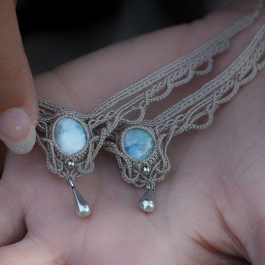 Duo de Bracelets de cheville bohème, ambiance plage et été sur les îles. Bijoux de pieds assortis. Micro macramé en fil de coton couleur lin beige avec une pierre de larimar bleu ciel accompagné d'une perle goutte argenté. Système de réglage à coulisse à l'arrière. Bijoux posé sur 2 mains dans un univers féérique bohème dans un jardin. Modèle avec Dreadlocks brunes, peau caramel et habits assorti au bijou de couleur lin naturel beige gris