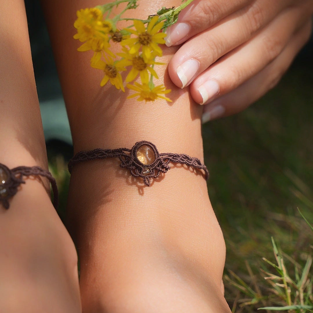 Bijoux de pieds, paire assortie de bracelets de chevilles à ambiance bohème celtique elfique. Fil marron foncé fin tissé en micro macramé avec entrelacs et arabesques, pierre centrale ovale petite taille jaune miel translucide. Bijoux hippie, bohème avec inspiration nature. Chevillères portées sur des chevilles à la peau pâle, bijoux de face avec jeu d'ombre et lumière et fleurs jaune en haut à droite.