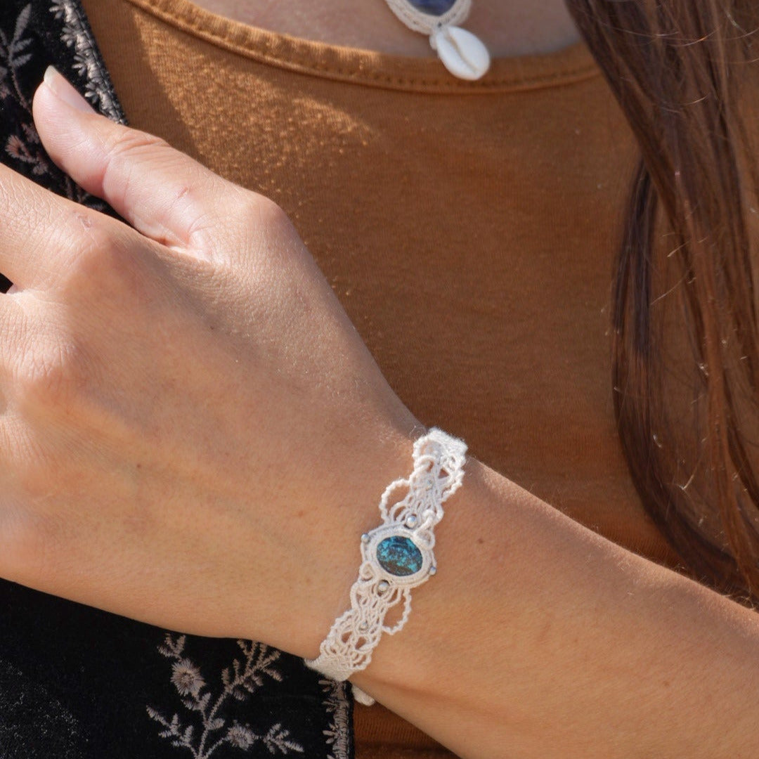 Bracelet large asymétrique. Bijou dentelle ambiance bohème hippie. Bracelet tissé en micro macramé avec fil de coton écru de 0.5 mm avec une pierre ovale de chrysocolle couleur bleu turquoise et marron foncé chocolat. Présence de part et d'autre de façon symétrique des perles en acier inoxydable argentées. Système de réglage à coulisse tubulaire à l'arrière. Design en relief et en torsade entrelacées celtiques. Ambiance plages, festival d'été, tribal. Bijou porté sur tenue bohème baroudeur