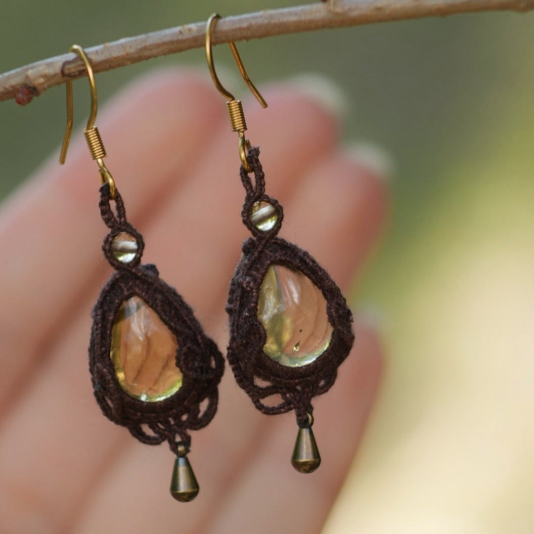 Boucles d'oreilles de style celtique elfique. Bijou féminin composé de : un tissage en relief avec arabesques en micro macramé avec du fil de coton marron foncé chocolat, pierre naturelle de citrine de forme goutte couleur jaune miel accompagné d'une perle ronde en citrine de 3 mm, d'un crochet doré en acier et d'une micro goutte bronze. Le bijou est présenté suspendu à une branche de noisetier avec en fond un main et du vert forestier naturel