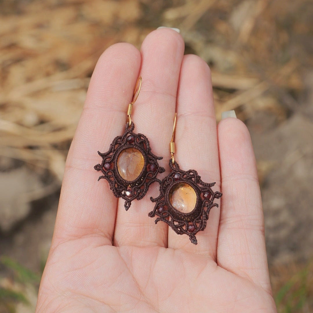 Paire de boucles d'oreilles et d'un collier ras-de-cou elfiques en micro macramé avec pierre naturelle. Bijoux tissé en fil de coton de petit diamètre. Mini Grenats rouge rosé facettés autour d'une citrine jaune ambré, fil de coton marron foncé. Motifs tissés en entrelacs celtiques et arabesques elfiques. Crochet doré. Bijoux ambiance bohème hippie aux couleurs chaudes. Bijoux posés, vue d'en haut dans une main à la peau claire.