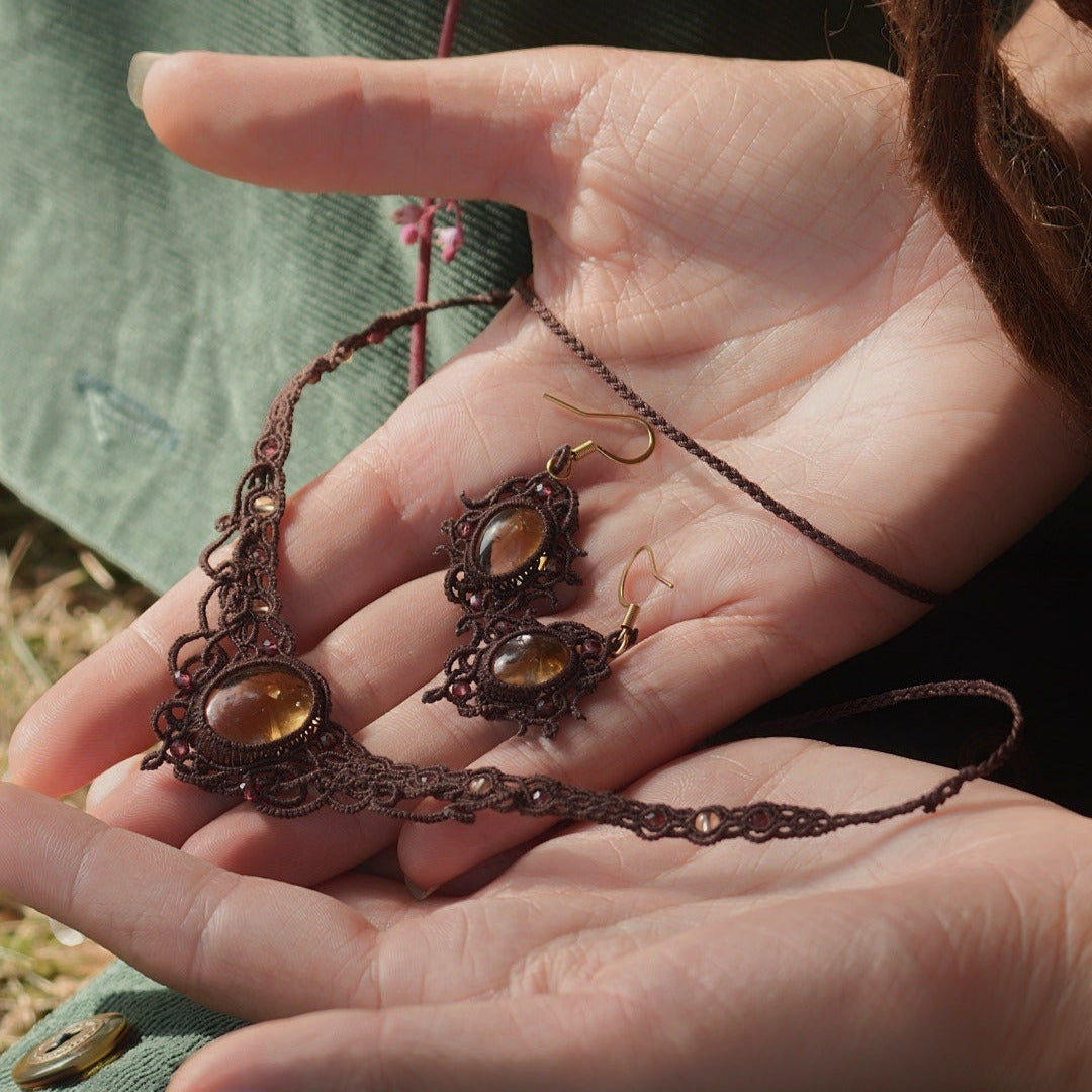 Parure de bijou composée d'une paire de boucles d'oreilles et d'un collier ras-de-cou elfiques en micro macramé avec pierre naturelle. Bijoux tissé en fil de coton de petit diamètre. Mini Grenats rouge rosé facettés autour d'une citrine jaune ambré, fil de coton marron foncé. Motifs tissés en entrelacs celtiques et arabesques elfiques. Crochet doré. Bijoux ambiance bohème hippie aux couleurs chaudes. Bijoux posés, vue d'en haut dans une main à la peau claire.