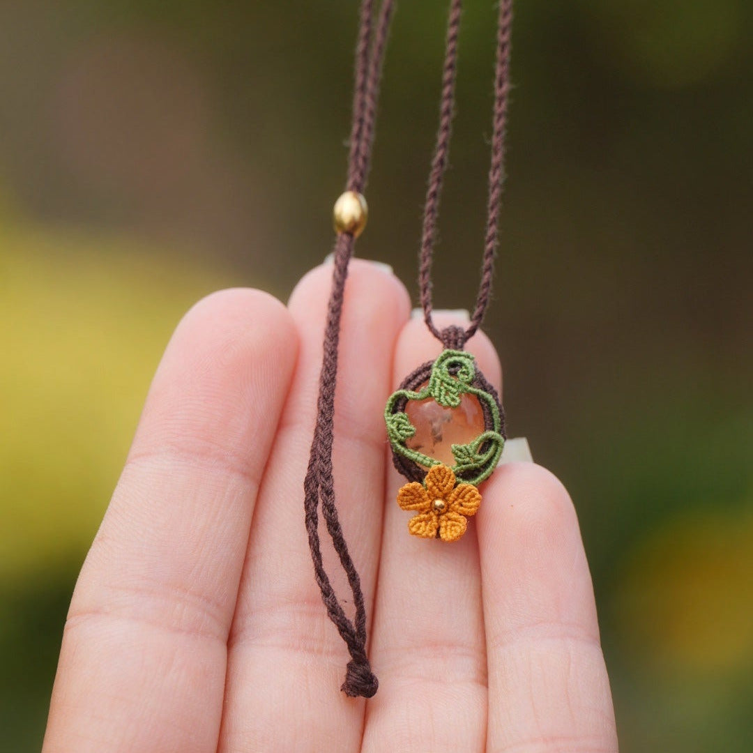 Collier avec pendentif en micro macramé avec motifs floraux. Bijou féminin composé d'un citrine jaune pâle translucide ovale sertie dans une coque marron foncé tissé avec un fil de 0.5 mm sur lequel est fixé une liane verte entrelacée avec mini feuilles et une fleur à 5 pétales jaune orangé avec un cœur doré de 2 mm.
Le bijou est en verticale, tenu dans le vide. le bijou est pris de face avec une main à la peau claire derrière