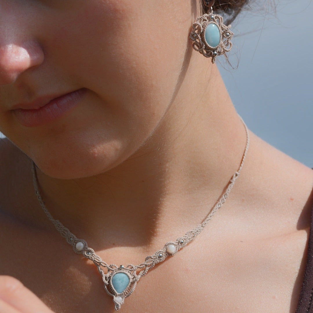 Parure Trésors d'Ecume composée d'une paire de boucles d'oreilles pendentes avec collier ras de cou assorti. Larimar bleu ciel ovales pour les boucles et en poire pointe en bas pour le collier. 3 Perles en nacre blanche pour le collier. Perles acier inoxydable argenté pour les deux pour le côté luxe et précieux. Dormeuses en acier inoxydable pour un port assuré. Fil de Coton couleur lin naturel (beige gris) de 0, 5 mm tissé en micro-macramé en relief version celtique. Bijoux porté femme brune peau caramel