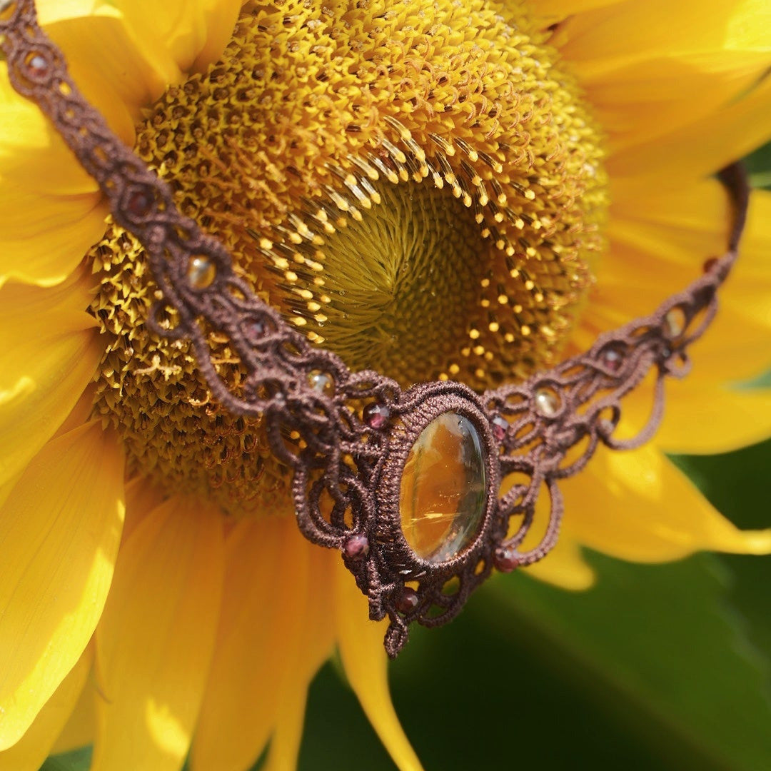 Collier ras-de-cou elfiques en micro macramé avec pierre naturelle. Bijoux tissé en fil de coton de petit diamètre. Mini Grenats rouge rosé facettés autour d'une citrine jaune ambré ovale, fil de coton marron foncé. Motifs tissés en entrelacs celtiques et arabesques elfiques Forme V. Bijoux ambiance bohème hippie aux couleurs chaudes. Bijoux posé à la verticale sur une fleur de tournesol jaune et nature. Bijoux vue de 3/4