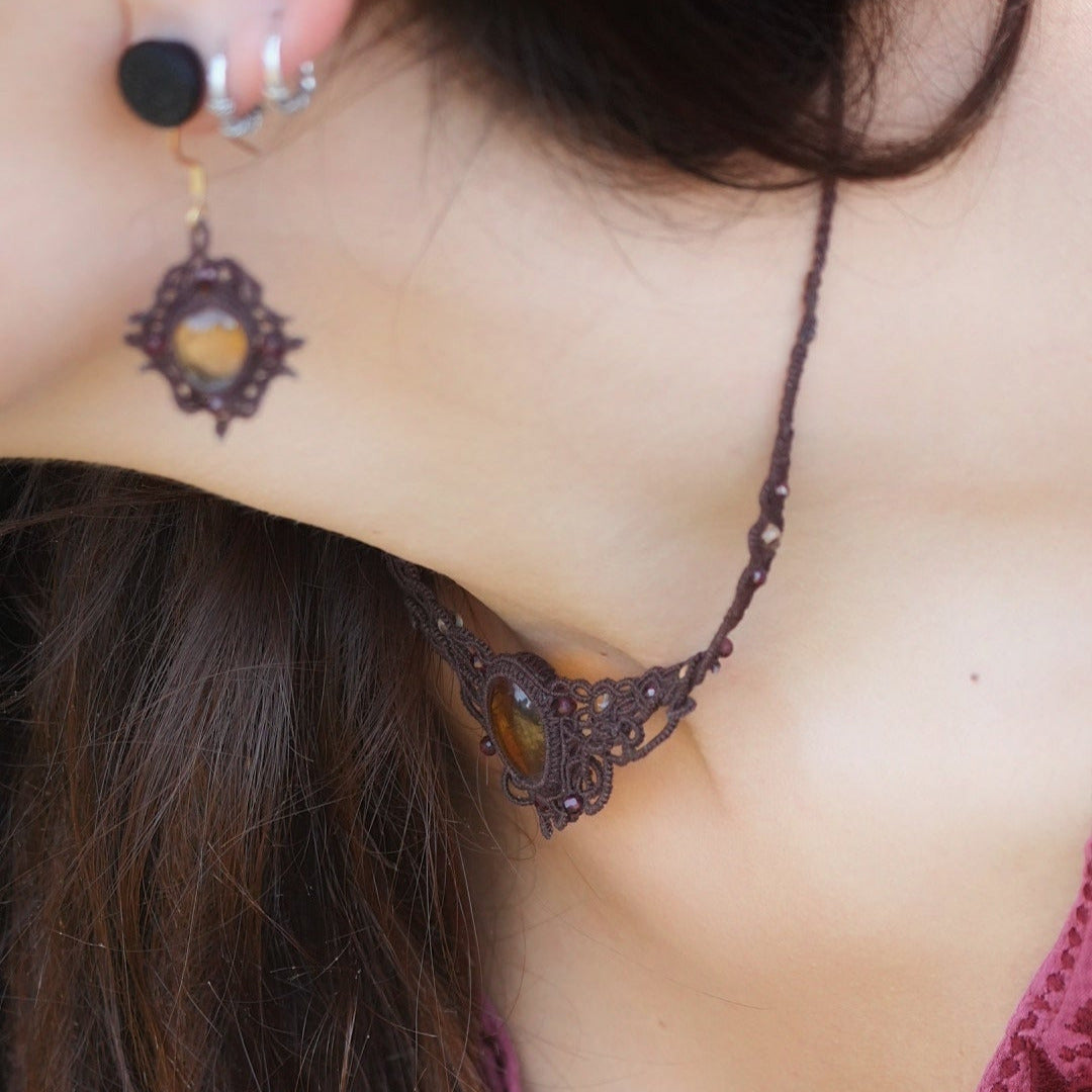 Collier ras-de-cou elfiques en micro macramé avec pierre naturelle. Bijoux tissé en fil de coton de petit diamètre. Mini Grenats rouge rosé facettés autour d'une citrine jaune ambré ovale, fil de coton marron foncé. Motifs tissés en entrelacs celtiques et arabesques elfiques Forme V. Bijoux ambiance bohème hippie aux couleurs chaudes. Bijoux porté sur un mannequin féminin à la eau claire et cheveux bruns, habit rose fuchsia. Vue de côté