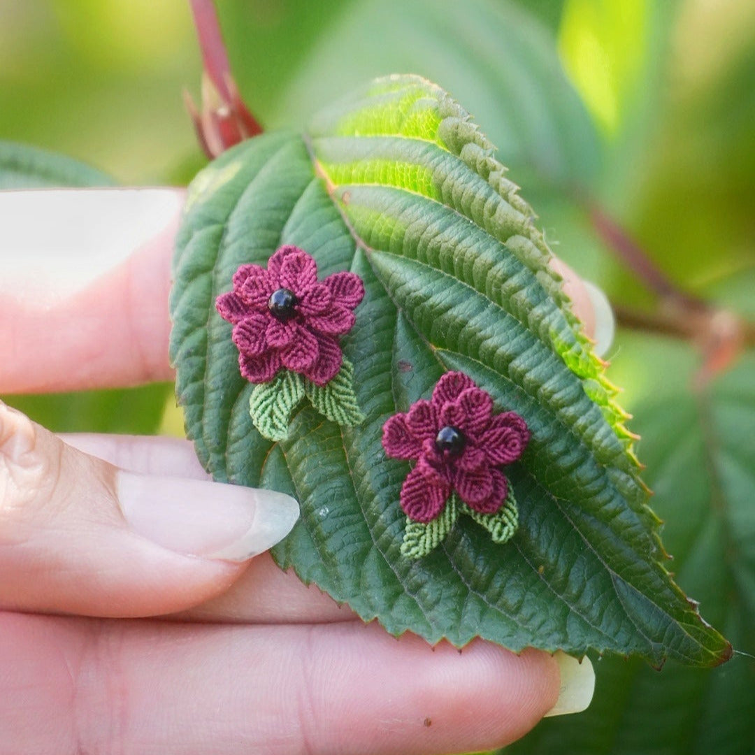 Boucles d’oreilles florales en micro macramé faites main, collection « Pétales éternels », posées sur une main féminine. Fleur 3D tissée en coton rouge bordeaux et vert tendre avec 13 pétales, 2 feuilles et cœur en perle d’onyx noire. Style bohème, elfique et hippie, idéal pour festivals et costumes féériques, photographié au milieu de feuilles et de nature verdoyante.