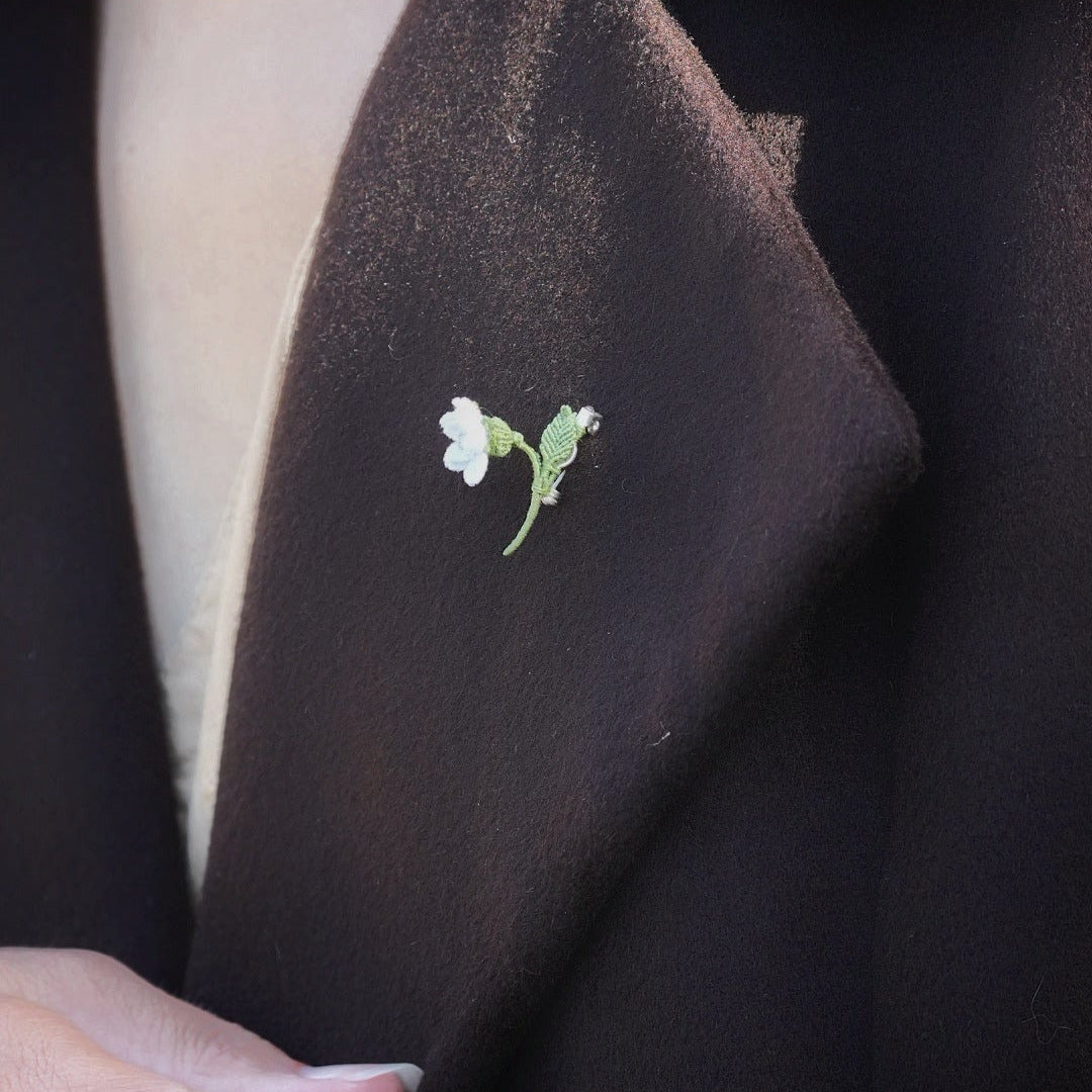 Mini broche fleur blanche en micro macramé avec cœur en labradorite bleu-vert, épinglée sur manteau en laine marron foncé, bijou elfique et bohème fait main.