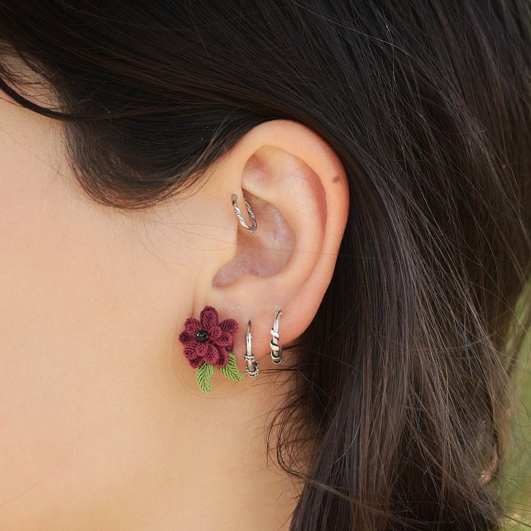 Boucles d’oreilles florales en micro macramé faites main, collection « Pétales éternels ». Bijou artisanal pour femme en coton rouge bordeaux et vert tendre, tissé en fleur 3D avec 13 pétales, 2 feuilles et un cœur en perle d’onyx noire de 3 mm. Fixées sur clous d’oreilles hypoallergéniques en acier inoxydable argenté avec poussoirs papillons. Style bohème, elfique et hippie, idéal pour festivals et costumes féériques. Photo portée sur modèle femme à peau claire et cheveux bruns.