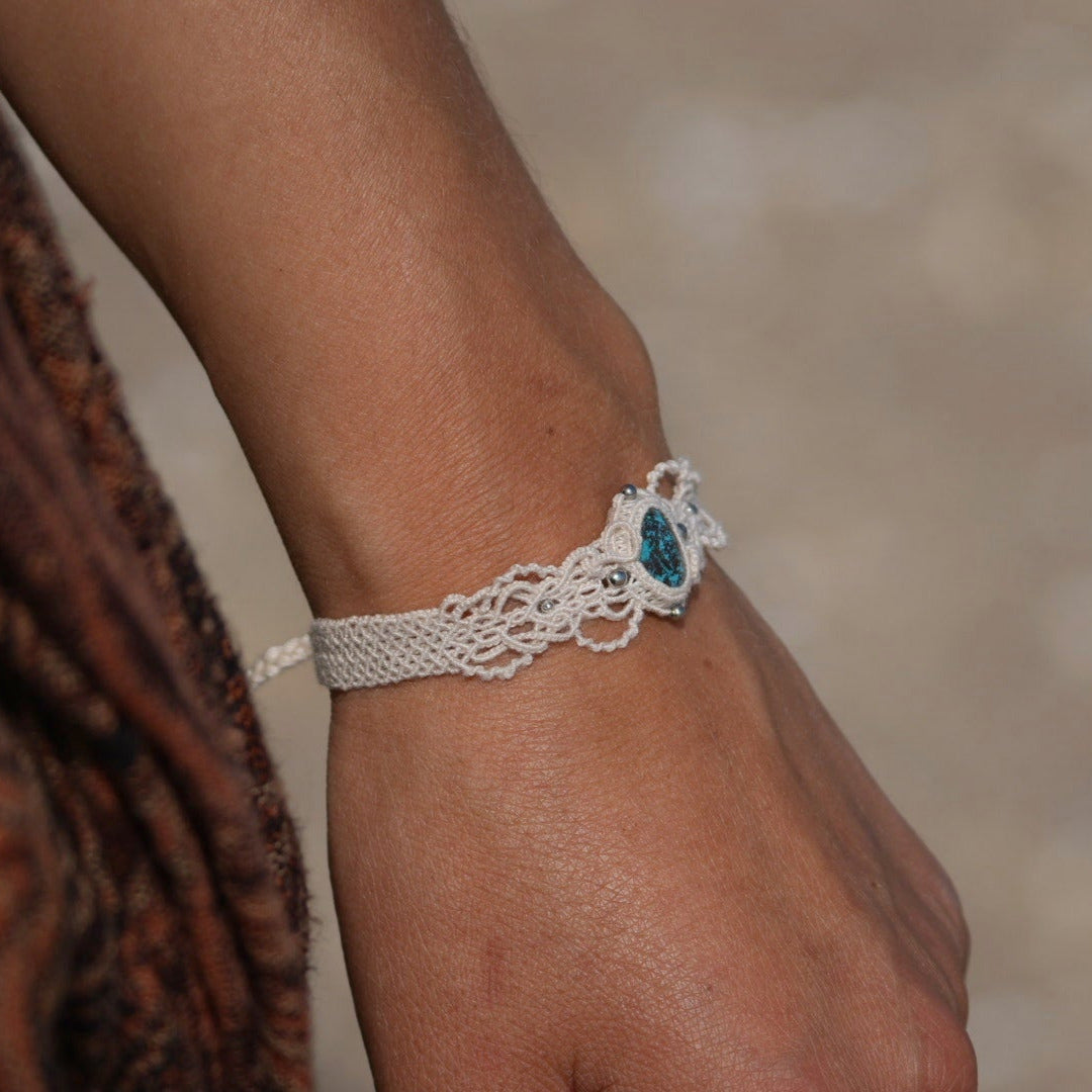 Bracelet large asymétrique. Bijou dentelle ambiance bohème hippie. Bracelet tissé en micro macramé avec fil de coton écru de 0.5 mm avec une pierre ovale de chrysocolle couleur bleu turquoise et marron foncé chocolat. Présence de part et d'autre de façon symétrique des perles en acier inoxydable argentées. Système de réglage à coulisse tubulaire à l'arrière. Design en relief et en torsade entrelacées celtiques. Ambiance plages, festival d'été, tribal. Bijou porté sur tenue bohème baroudeur
