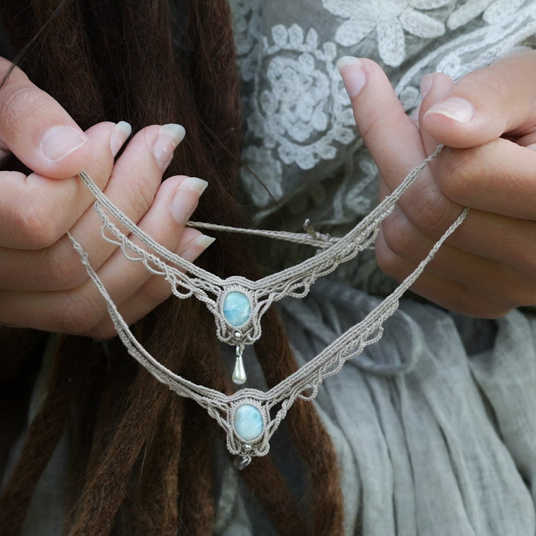 Duo de Bracelets de cheville bohème, ambiance plage et été sur les îles. Bijoux de pieds assortis. Micro macramé en fil de coton couleur lin beige avec une pierre de larimar bleu ciel accompagné d'une perle goutte argenté. Système de réglage à coulisse à l'arrière. Bijoux tenus par 2 mains dans un univers féérique bohème dans un jardin. Modèle avec Dreadlocks brunes, peau caramel et habits assorti au bijou de couleur lin naturel beige gris