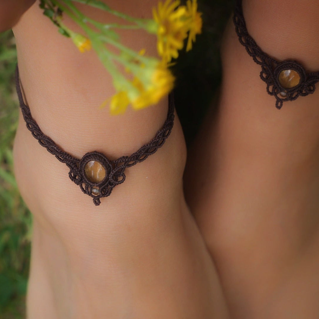 Bijoux de pieds, paire assortie de bracelets de chevilles à ambiance bohème celtique elfique. Fil marron foncé fin tissé en micro macramé avec entrelacs et arabesques, pierre centrale ovale petite taille jaune miel translucide. Bijoux hippie, bohème avec inspiration nature. Chevillères portées sur des chevilles à la peau pâle, bijoux de face avec jeu d'ombre et lumière et fleurs jaune en haut à droite.
