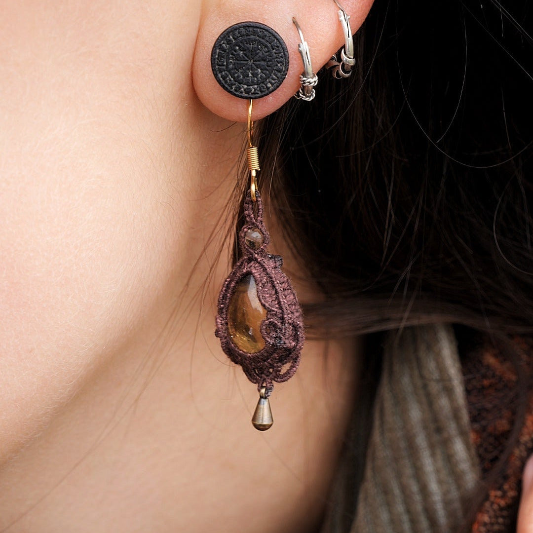 Boucles d'oreilles de style celtique elfique. Bijou féminin composé de : un tissage en relief avec arabesques en micro macramé avec du fil de coton marron foncé chocolat, pierre naturelle de citrine de forme goutte couleur jaune miel accompagné d'une perle ronde en citrine de 3 mm, d'un crochet doré en acier et d'une micro goutte bronze. Le bijou est porté sur une peau dorée et avec un fond de cheveux brun foncé, une main dans le cou avec une écharper aux tons orangers