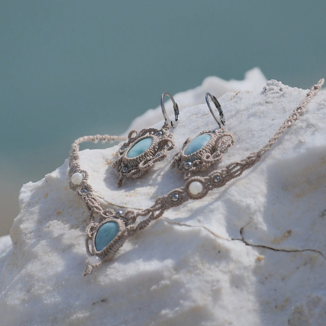 Parure Trésors d'Ecume composée d'une paire de boucles d'oreilles pendentes avec collier ras de cou assorti. Larimar bleu ciel ovales pour les boucles et en poire pointe en bas pour le collier. 3 Perles en nacre blanche pour le collier. Perles acier inoxydable argenté pour les deux pour le côté luxe et précieux. Dormeuses en acier inoxydable pour un port assuré. Fil de Coton couleur lin naturel (beige gris) de 0, 5 mm tissé en micro-macramé en relief version celtique. Bijoux posés sur une pierre blanche