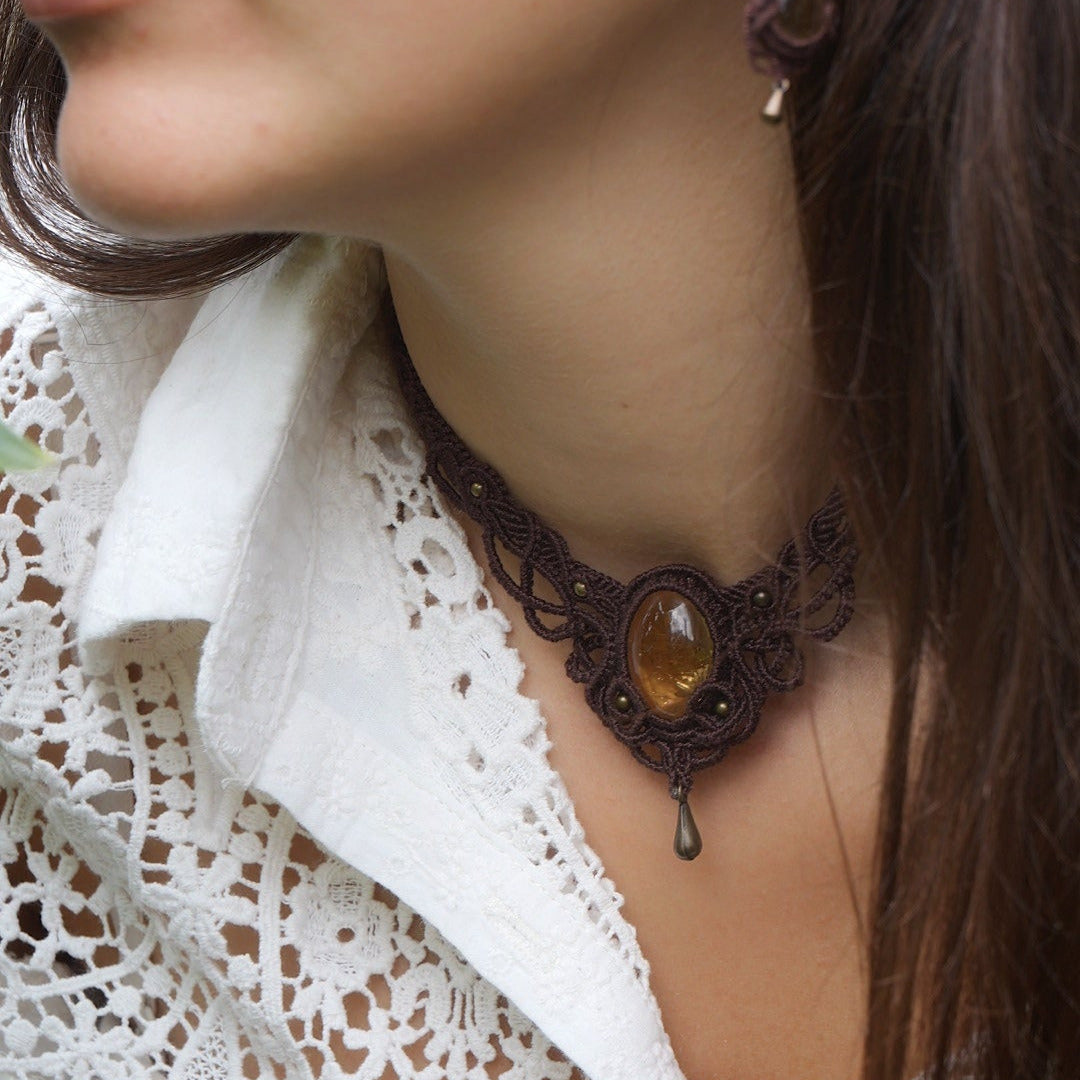Collier large en micro macramé de couleur marron foncé, jaune ambré et bronze. Motif tissé en micro macramé avec ambiance entrelacs celtiques et arabesques elfiques. Présence d'une pierre centrale ovale de grande taille jaune translucide, gouttes bronze, forme V avec mini perles ronde bronze parsemées. Photo ambiance bohème hippie avec une touche d'élégance. Bijou porté sur une peau claire avec chemise blanche en dentelle.