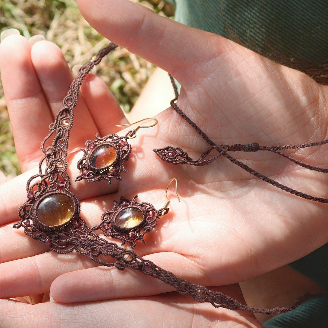 Parure de bijou composée d'une paire de boucles d'oreilles et d'un collier ras-de-cou elfiques en micro macramé avec pierre naturelle. Bijoux tissé en fil de coton de petit diamètre. Mini Grenats rouge rosé facettés autour d'une citrine jaune ambré, fil de coton marron foncé. Motifs tissés en entrelacs celtiques et arabesques elfiques. Crochet doré. Bijoux ambiance bohème hippie aux couleurs chaudes. Bijoux posés, vue d'en haut dans une main à la peau claire.
