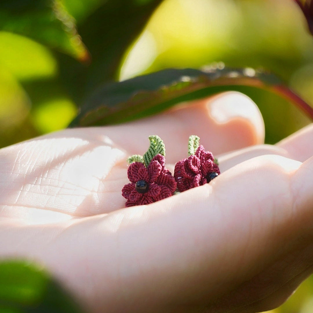 Boucles d’oreilles florales en micro macramé faites main, collection « Pétales éternels », posées sur une main féminine. Fleur 3D tissée en coton rouge bordeaux et vert tendre avec 13 pétales, 2 feuilles et cœur en perle d’onyx noire. Style bohème, elfique et hippie, idéal pour festivals et costumes féériques, photographié au milieu de feuilles et de nature verdoyante.