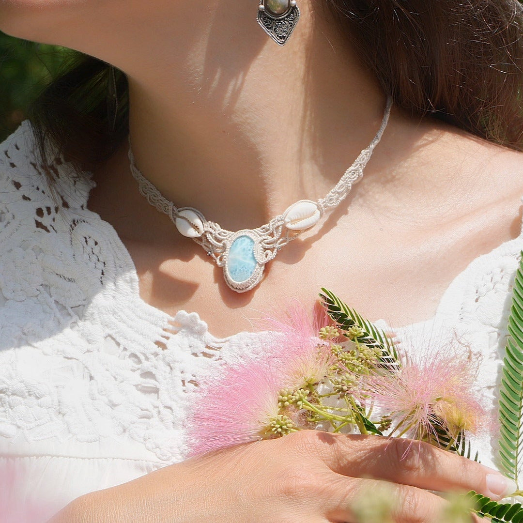 Collier plastron en Larimar Océan bleu ciel, coton couleur lin naturel (beige gris), cauris blancs et perles en acier inoxydable argenté. Système de réglable en tube tissé à l'arrière du bijou. Tissage symétrique en micro macramé avec effet nœuds celtiques entrelacés en relief. Style bohème, hippie, plage, Sirène, tribal et tropical. Convient pour un mariage type bohème ou plage. Bijou porté avec une tenue romantique et bohème blanche avec dentelle apparente. Modèle à la peau bronzée et aux cheveux bruns.