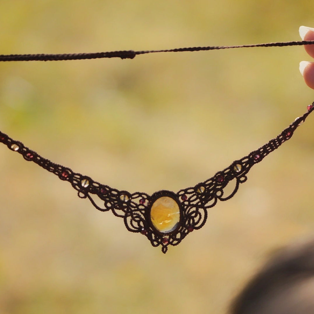 Collier ras-de-cou elfiques en micro macramé avec pierre naturelle. Bijoux tissé en fil de coton de petit diamètre. Mini Grenats rouge rosé facettés autour d'une citrine jaune ambré ovale, fil de coton marron foncé. Motifs tissés en entrelacs celtiques et arabesques elfiques Forme V. Bijoux ambiance bohème hippie aux couleurs chaudes. Bijoux tenu à la verticale avec fond claire et nature. Bijoux vue de face avec transparence et jeu de lumière