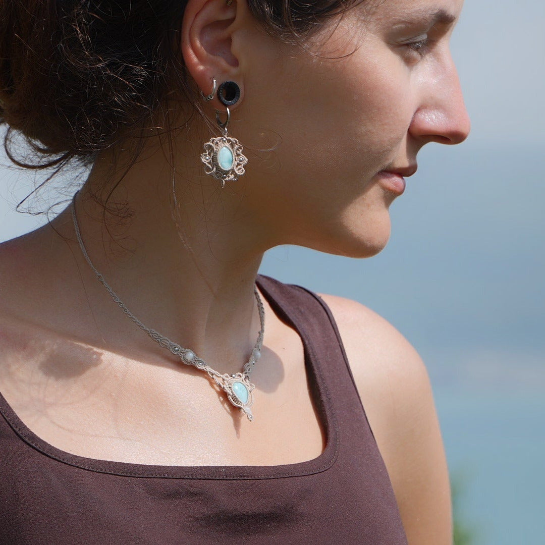 Parure Trésors d'Ecume composée d'une paire de boucles d'oreilles pendentes avec collier ras de cou assorti. Larimar bleu ciel ovales pour les boucles et en poire pointe en bas pour le collier. 3 Perles en nacre blanche pour le collier. Perles acier inoxydable argenté pour les deux pour le côté luxe et précieux. Dormeuses en acier inoxydable pour un port assuré. Fil de Coton couleur lin naturel (beige gris) de 0, 5 mm tissé en micro-macramé en relief version celtique. Bijoux porté femme brune peau caramel