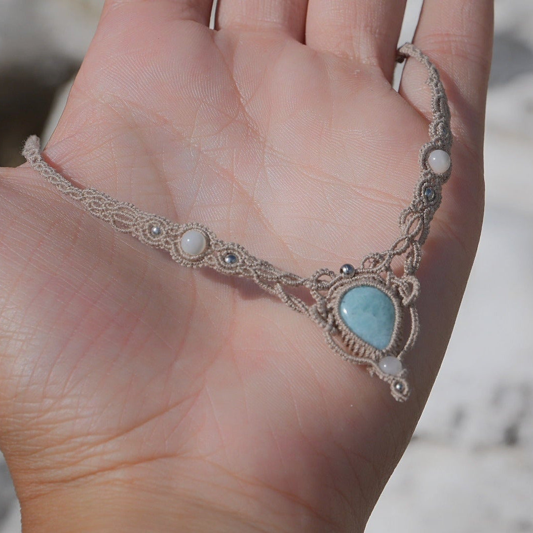Collier ras-de-cou forme v Trésors d'Ecume composée d'une Larimar bleu ciel en poire pointe en bas, perles en nacre blanche ronde rappel des coquillages et de la plage, mini Perles rondes en acier inoxydable argenté pour le côté luxe et précieux. Fil de Coton couleur lin naturel (beige gris) de 0, 5 mm tissé en micro-macramé en relief version celtique. Collage 3 photos : Bijoux à plat dans une main à la peau claire. Rocher blanc en fond. . Photo ambiance tropiques et plages, style bohème hippie