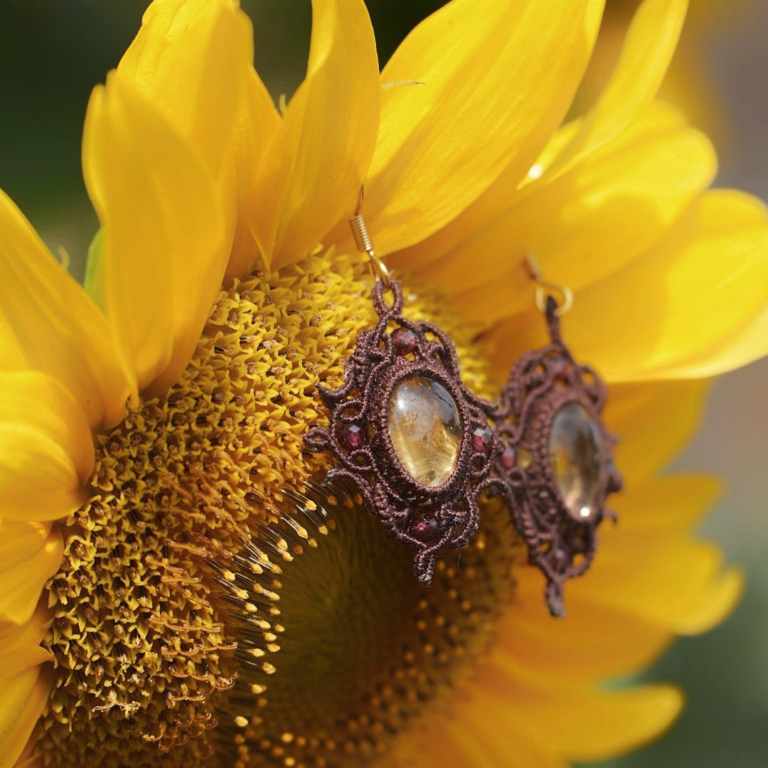 Boucles d'oreilles elfiques en micro macramé avec pierre naturelle. Bijoux tissé en fil de coton de petit diamètre. 4 Mini Grenats rouge rosé facettés autour d'une citrine jaune ambré, fil de coton marron foncé. Motifs tissés en entrelacs celtiques et arabesques elfiques. Crochet doré. Bijou ambiance bohème hippie aux couleurs chaudes. Bijou posé à la verticale, vue de 3/4 sur un tournesol jaune