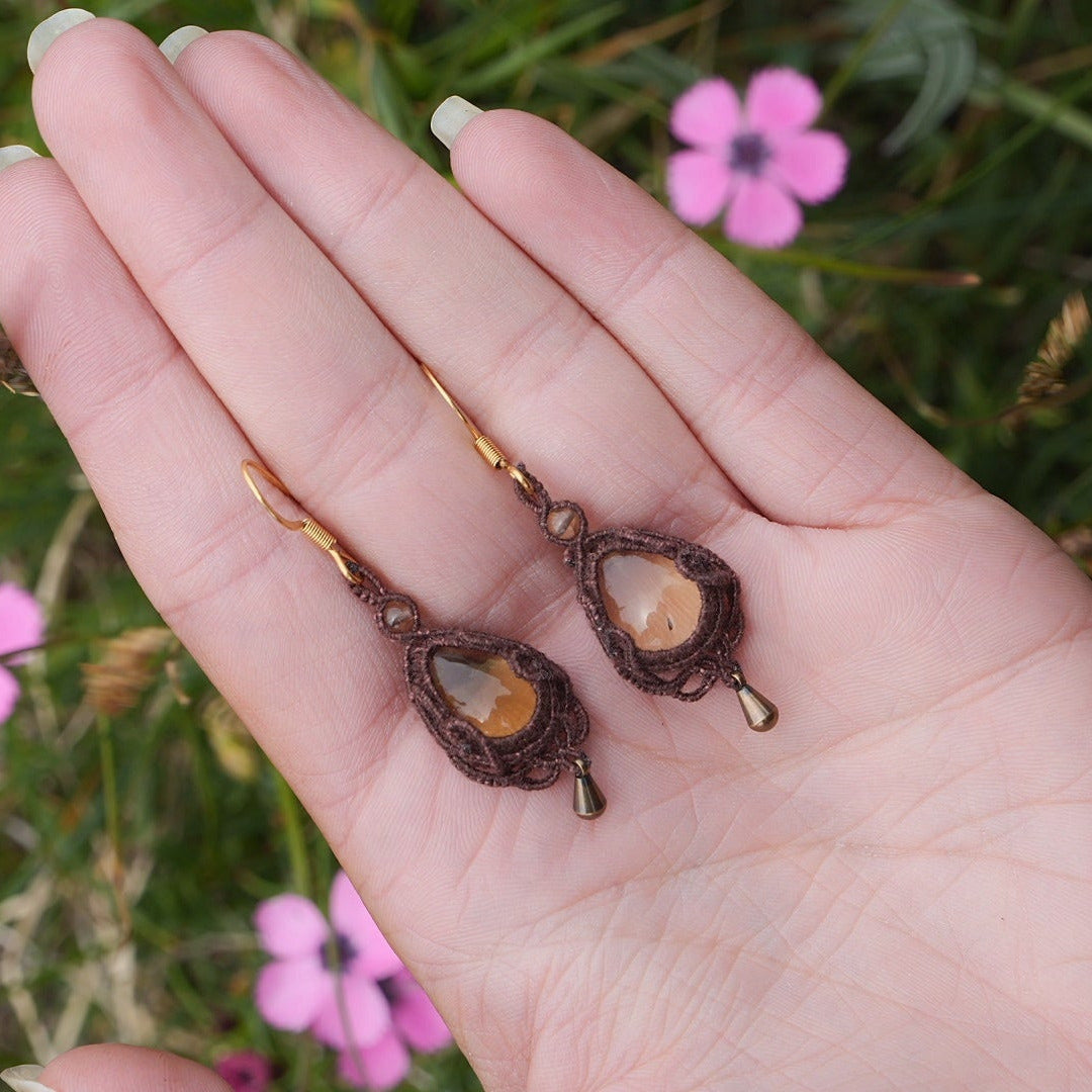 Boucles d'oreilles de style celtique elfique. Bijou féminin composé de : un tissage en relief avec arabesques en micro macramé avec du fil de coton marron foncé chocolat, pierre naturelle de citrine de forme goutte couleur jaune miel accompagné d'une perle ronde en citrine de 3 mm, d'un crochet doré en acier et d'une micro goutte bronze. Le bijou est posé dans une main pâle avec un fond nature vert accompagné d’œillet rose