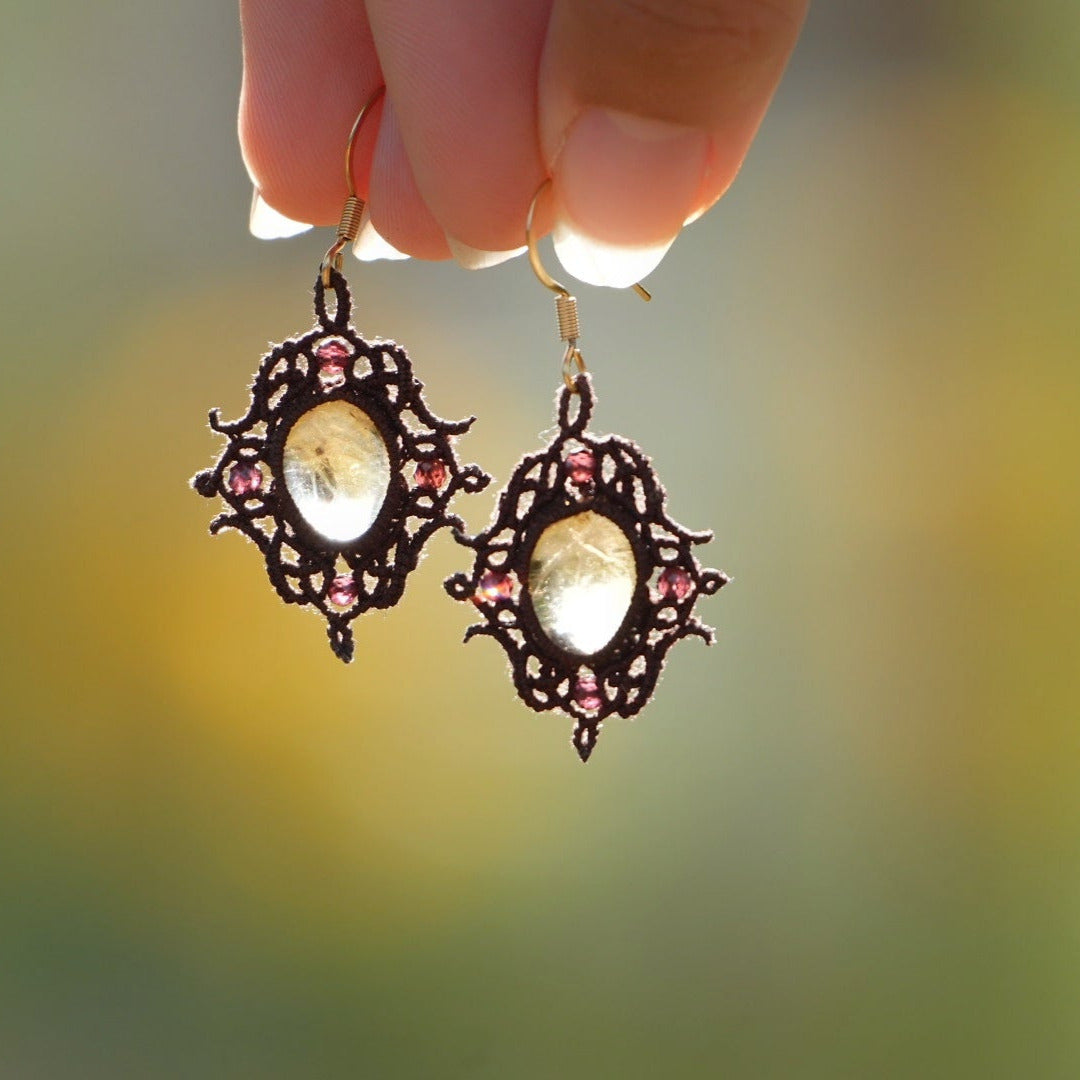 Boucles d'oreilles elfiques en micro macramé avec pierre naturelle. Bijoux tissé en fil de coton de petit diamètre. 4 Mini Grenats rouge rosé facettés translucides autour d'une citrine jaune ambré translucide, fil de coton marron foncé. Motifs tissés en entrelacs celtiques et arabesques elfiques. Crochet doré. Bijou ambiance bohème hippie aux couleurs chaudes. Bijou tenu dans une mains de façon verticale, prise de vue de face avec un jeu de lumière de façon contrejour