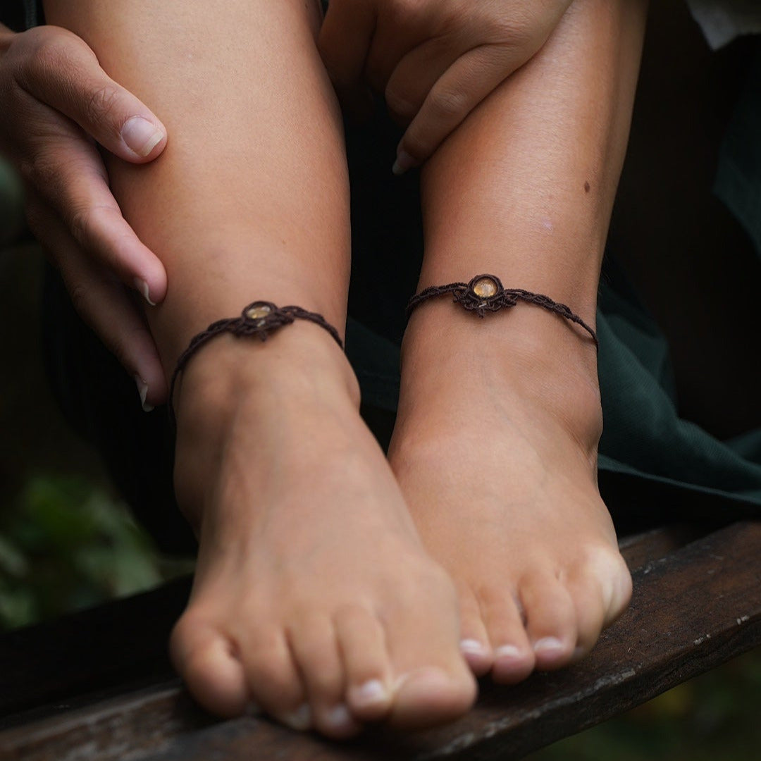 Bijoux de pieds, paire assortie de bracelets de chevilles à ambiance bohème celtique elfique. Fil marron foncé fin tissé en micro macramé avec entrelacs et arabesques, pierre centrale ovale petite taille jaune miel translucide. Bijoux hippie, bohème avec inspiration nature. Chevillères portées sur des chevilles à la peau pâle, ambiance sombre/dark. Modèle assise sur un banc en bois, les jambes relevées, les mains autour des mollets, de face avec jeu d'ombre en fond, jupe velours côtelé vert sapin