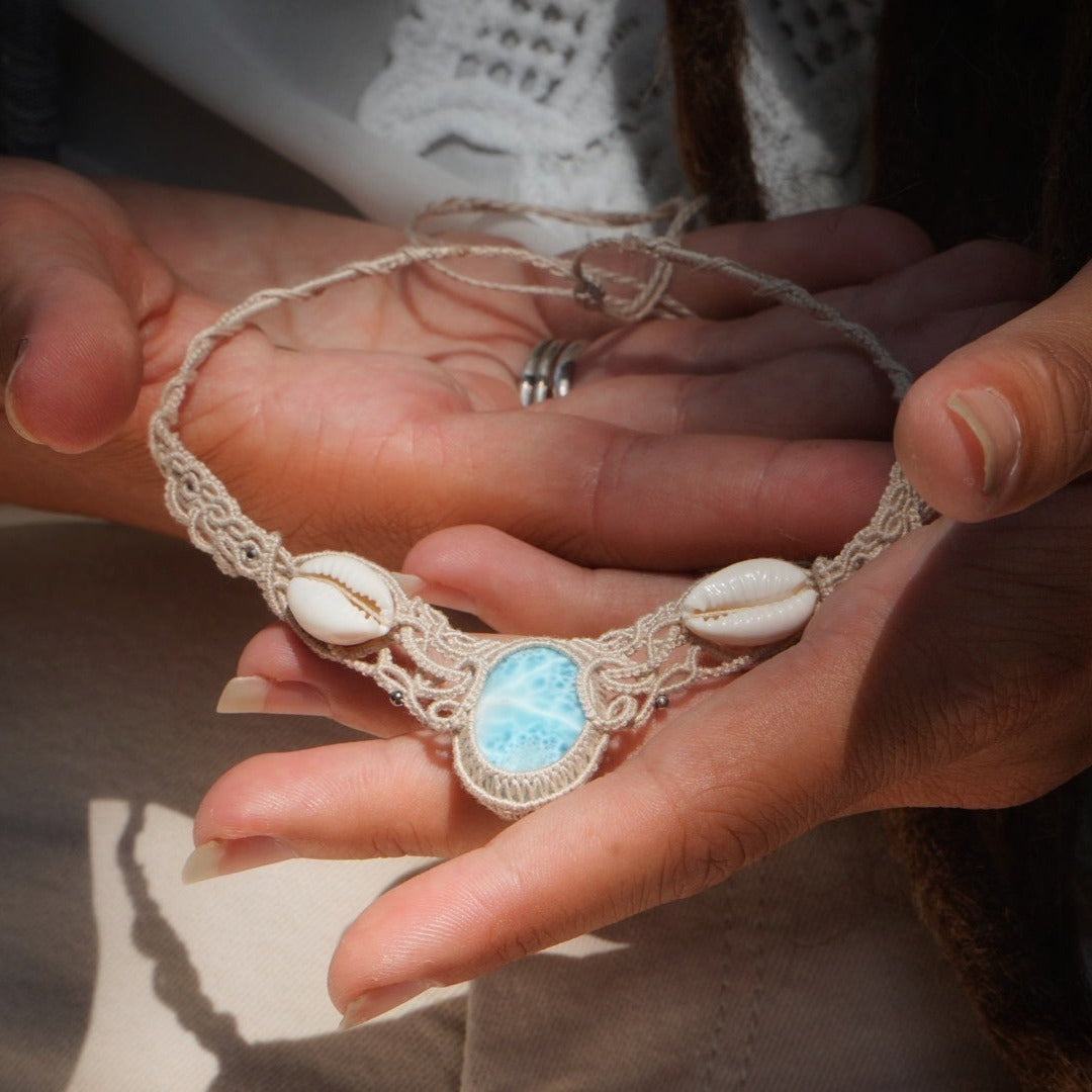 Collier plastron en Larimar Océan bleu ciel, coton couleur lin naturel (beige gris), cauris blancs et perles en acier inoxydable argenté. Système de réglable en tube tissé à l'arrière du bijou. Tissage symétrique en micro macramé avec effet nœuds celtiques entrelacés en relief. Style bohème, hippie, plage, Sirène, tribal et tropical. Convient pour un mariage type bohème ou plage. Bijou tenu vue 3/4, tenue romantique et bohème blanche avec dentelle apparente. Modèle à la peau bronzée et aux cheveux bruns.