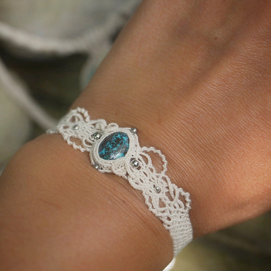 Bracelet large asymétrique. Bijou dentelle ambiance bohème hippie. Bracelet tissé en micro macramé avec fil de coton écru de 0.5 mm avec une pierre ovale de chrysocolle couleur bleu turquoise et marron foncé chocolat. Présence de part et d'autre de façon symétrique des perles en acier inoxydable argentées. Système de réglage à coulisse tubulaire à l'arrière. Design en relief et en torsade entrelacées celtiques. Ambiance plages, festival d'été, tribal. Bijou porté fond mer et ciel, pantalon beige lagre.