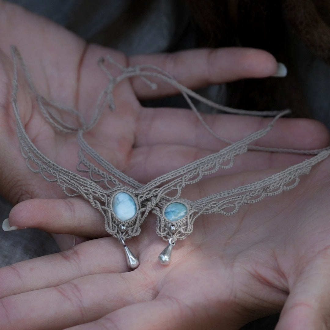 Duo de Bracelets de cheville bohème, ambiance plage et été sur les îles. Bijoux de pieds assortis. Micro macramé en fil de coton couleur lin beige avec une pierre de larimar bleu ciel accompagné d'une perle goutte argenté. Système de réglage à coulisse à l'arrière. Bijoux posé sur 2 mains dans un univers féérique bohème dans un jardin. Modèle avec Dreadlocks brunes, peau caramel et habits assorti au bijou de couleur lin naturel beige gris