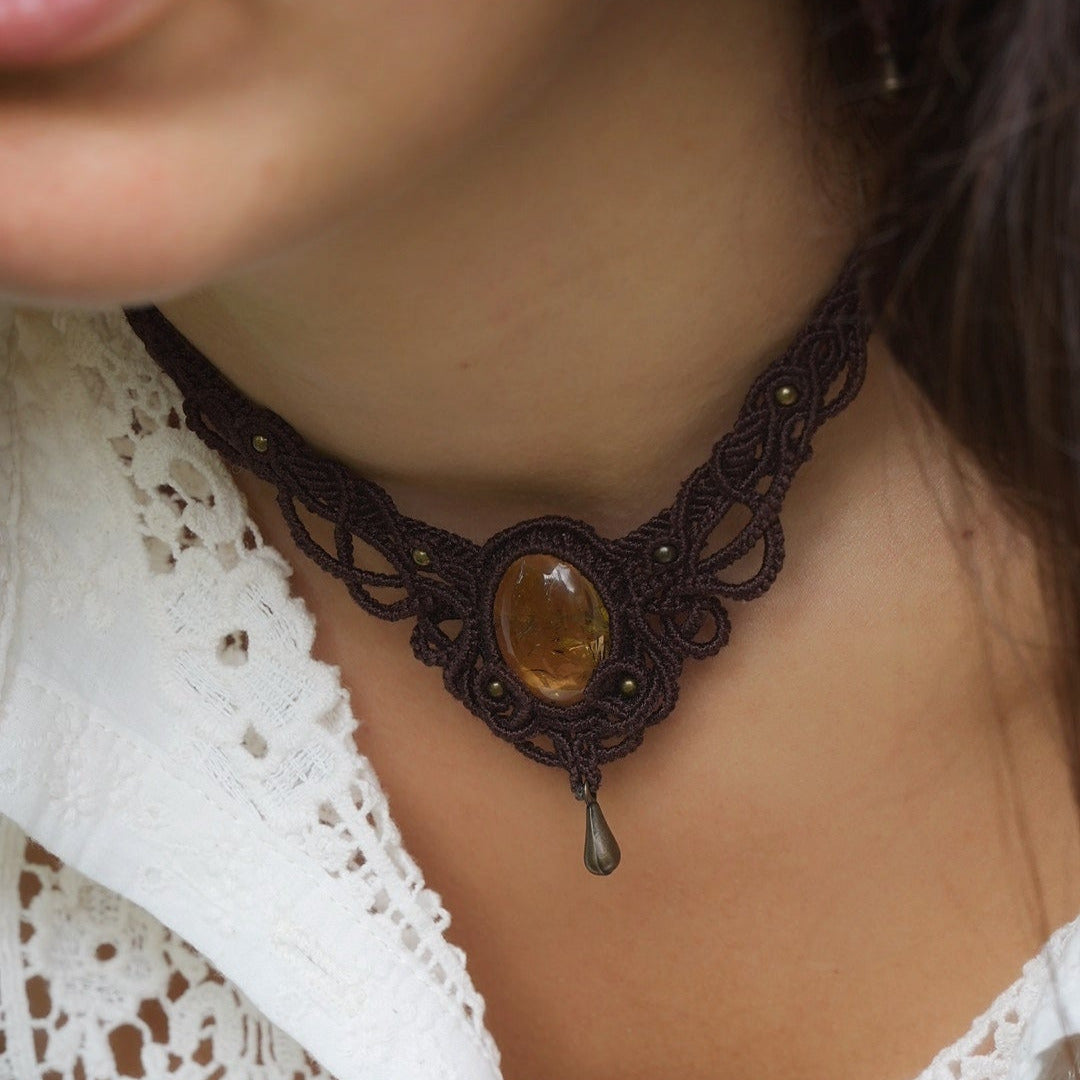 Collier large en micro macramé de couleur marron foncé, jaune ambré et bronze. Motif tissé en micro macramé avec ambiance entrelacs celtiques et arabesques elfiques. Présence d'une pierre centrale ovale de grande taille jaune translucide, gouttes bronze, forme V avec mini perles ronde bronze parsemées. Photo ambiance bohème hippie avec une touche d'élégance. Bijou porté sur une peau claire avec chemise blanche en dentelle.