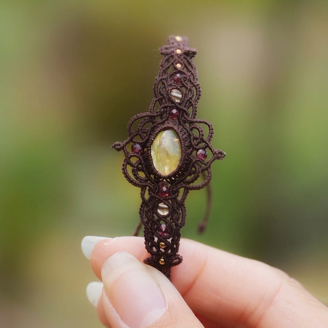 Bracelet réglable en micro macramé. Bijou féminin composé d'une citrine jaune ovale translucide au centre, entourée de 6 perles en grenat rouge à facettes de 3 mm puis de 3 perles dorées de 2 mm. Fil de coton de 0.4 mm tissé en micro macramé avec de motifs en reliefs type nœuds celtiques et motifs arabesques féériques et elfiques. Bijou large ambiance et style bohème hippie, fairycore.
Bijou tenu par une main de femme à la peau claire avec fond vert nature. Vue de face
