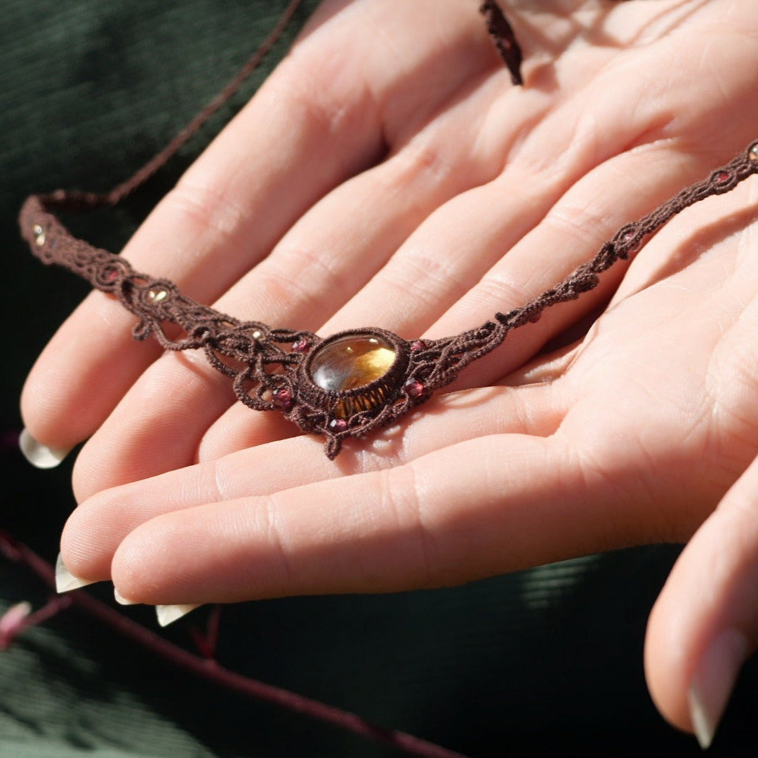 Collier ras-de-cou elfiques en micro macramé avec pierre naturelle. Bijoux tissé en fil de coton de petit diamètre. Mini Grenats rouge rosé facettés autour d'une citrine jaune ambré ovale, fil de coton marron foncé. Motifs tissés en entrelacs celtiques et arabesques elfiques Forme V. Bijoux ambiance bohème hippie aux couleurs chaudes. Bijoux posé à plat dans 2 mains en coupe à la eau claire et fond velours côtelé vert Vue de 3/4
