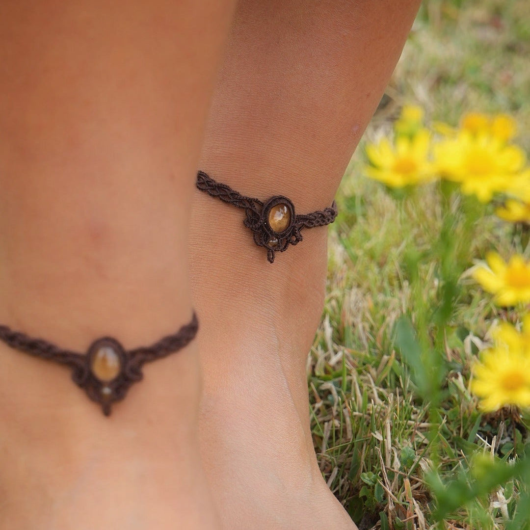 Bijoux de pieds, paire assortie de bracelets de chevilles à ambiance bohème celtique elfique. Fil marron foncé fin tissé en micro macramé avec entrelacs et arabesques, pierre centrale ovale petite taille jaune miel translucide. Bijoux hippie, bohème avec inspiration nature. Chevillères portées sur des chevilles à la peau pâle prise de face avec fleurs jaune à droite au premier plan et fond herbe verte.