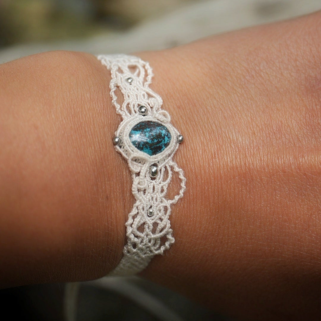 Bracelet large asymétrique. Bijou dentelle ambiance bohème hippie. Bracelet tissé en micro macramé avec fil de coton écru de 0.5 mm avec une pierre ovale de chrysocolle couleur bleu turquoise et marron foncé chocolat. Présence de part et d'autre de façon symétrique des perles en acier inoxydable argentées. Système de réglage à coulisse tubulaire à l'arrière. Design en relief et en torsade entrelacées celtiques. Ambiance plages, festival d'été, tribal. Bijou porté fond bois clair