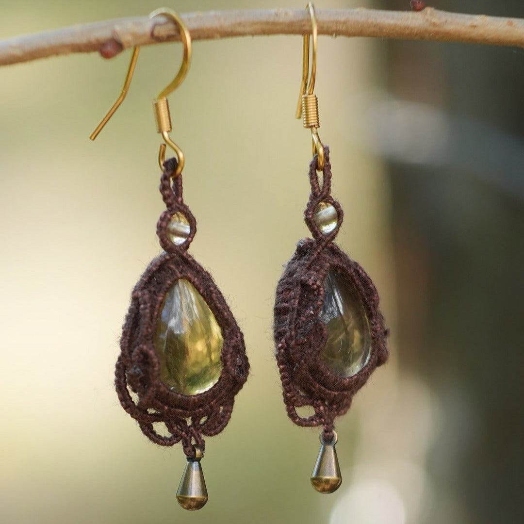 Boucles d'oreilles de style celtique elfique. Bijou féminin composé de : un tissage en relief avec arabesques en micro macramé avec du fil de coton marron foncé chocolat, pierre naturelle de citrine de forme goutte couleur jaune miel accompagné d'une perle ronde en citrine de 3 mm, d'un crochet doré en acier et d'une micro goutte bronze. Le bijou est présenté suspendu à une branche de noisetier avec un fond vert forestier naturel