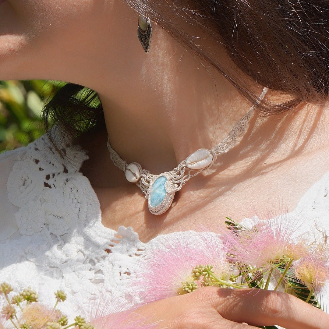 Collier plastron en Larimar Océan bleu ciel, coton couleur lin naturel (beige gris), cauris blancs et perles en acier inoxydable argenté. Système de réglable en tube tissé à l'arrière du bijou. Tissage symétrique en micro macramé avec effet nœuds celtiques entrelacés en relief. Style bohème, hippie, plage, Sirène, tribal et tropical. Convient pour un mariage type bohème ou plage. Bijou porté avec une tenue romantique et bohème blanche avec dentelle apparente. Modèle à la peau bronzée et aux cheveux bruns.