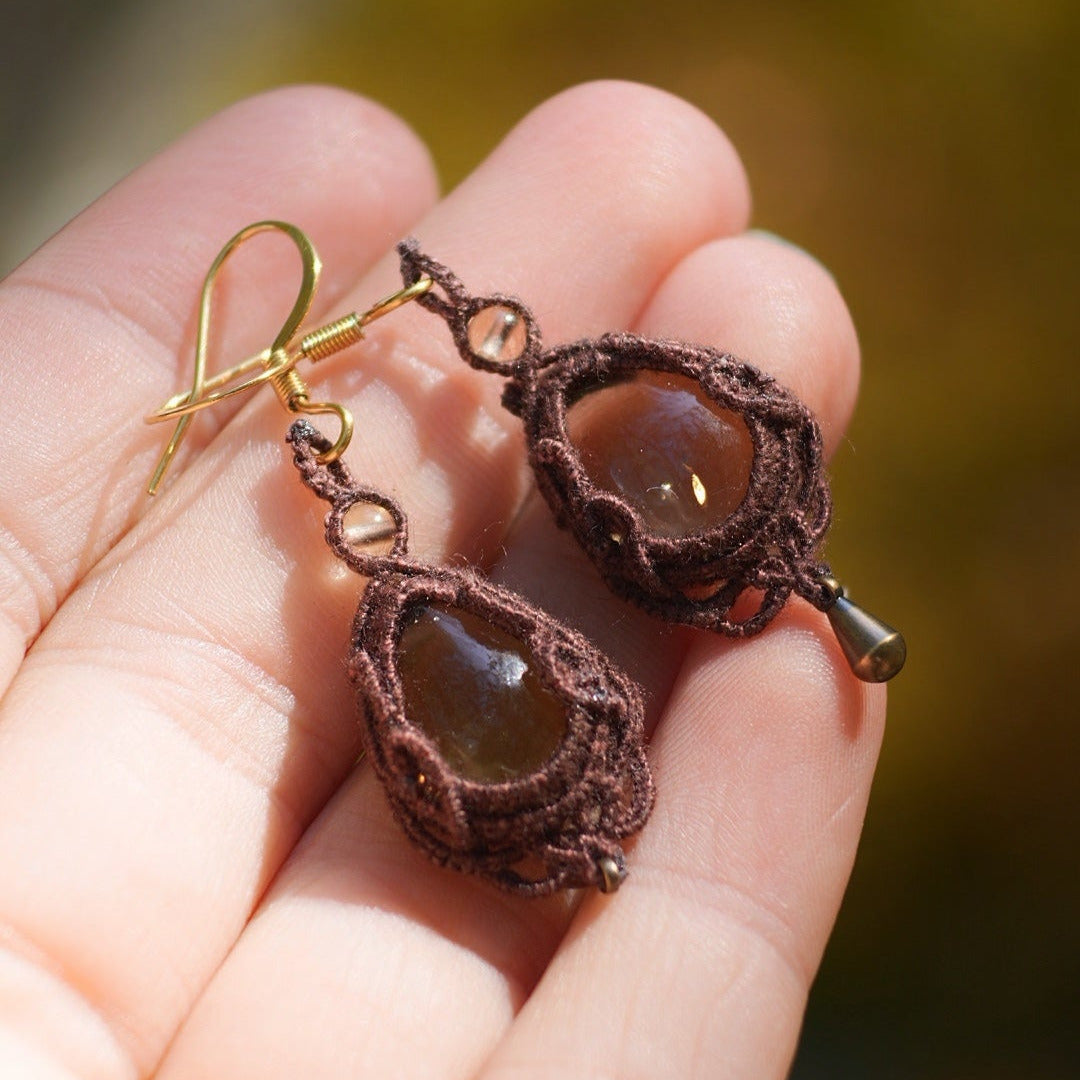 Boucles d'oreilles de style celtique elfique. Bijou féminin composé de : un tissage en relief avec arabesques en micro macramé avec du fil de coton marron foncé chocolat, pierre naturelle de citrine de forme goutte couleur jaune miel accompagné d'une perle ronde en citrine de 3 mm, d'un crochet doré en acier et d'une micro goutte bronze. Le bijou est posé dans une main pâle avec un fond nature vert jaune