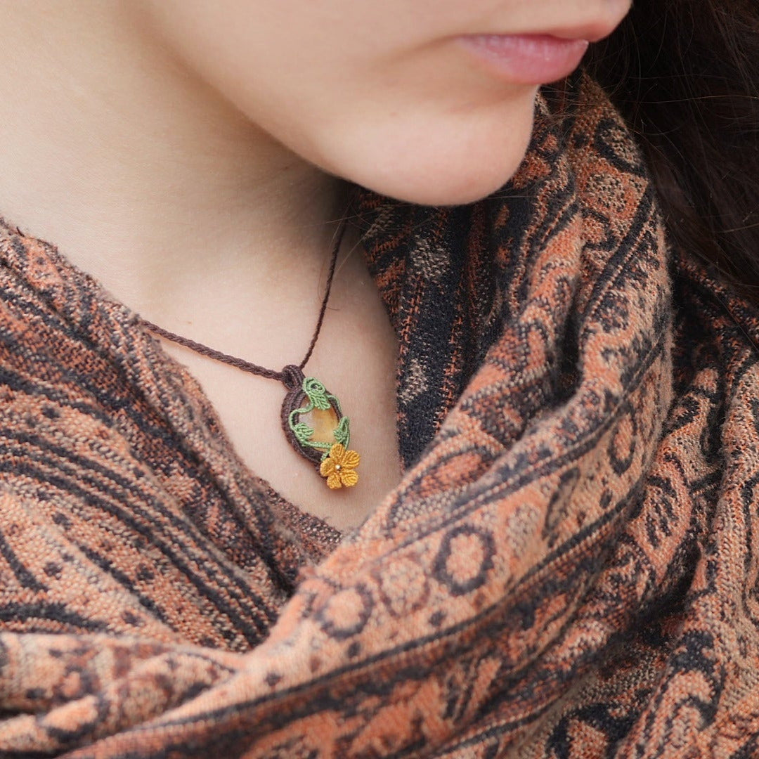 Collier avec pendentif en micro macramé avec motifs floraux. Bijou féminin composé d'un citrine jaune pâle translucide ovale sertie dans une coque marron foncé tissé avec un fil de 0.5 mm sur lequel est fixé une liane verte entrelacée avec mini feuilles et une fleur à 5 pétales jaune orangé avec un cœur doré de 2 mm.
Le bijou est porté autour d'un cou de femme à la peau claire et d'un châle orange et noir. le bijou est pris de 3/4