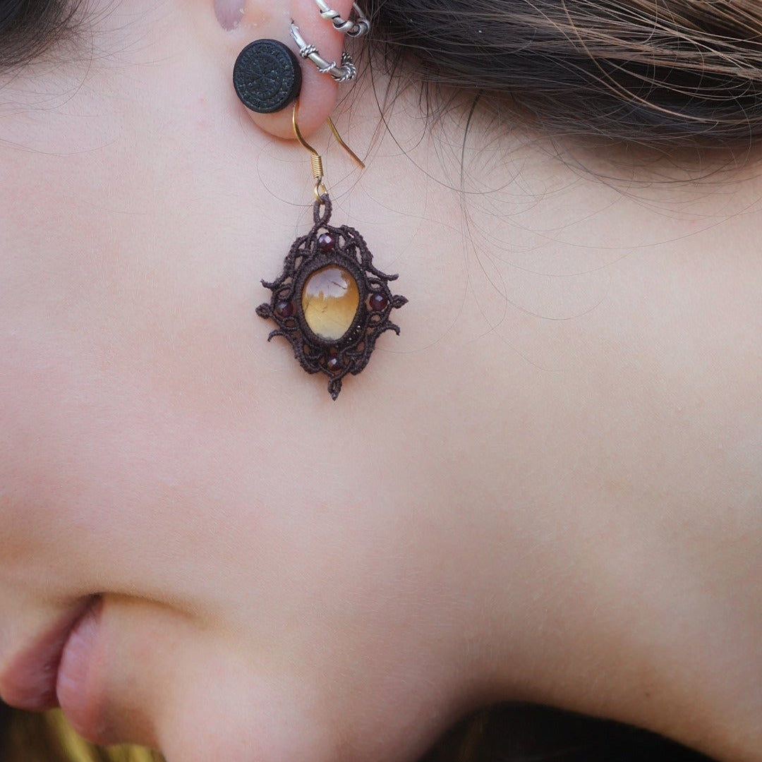 Boucles d'oreilles elfiques en micro macramé avec pierre naturelle. Bijoux tissé en fil de coton de petit diamètre. 4 Mini Grenats rouge rosé facettés autour d'une citrine jaune ambré, fil de coton marron foncé. Motifs tissés en entrelacs celtiques et arabesques elfiques. Crochet doré. Bijou ambiance bohème hippie aux couleurs chaudes. Bijou porté, vue de face sur un modèle féminin à la peau claire et aux cheveux bruns. Habits rose font vert