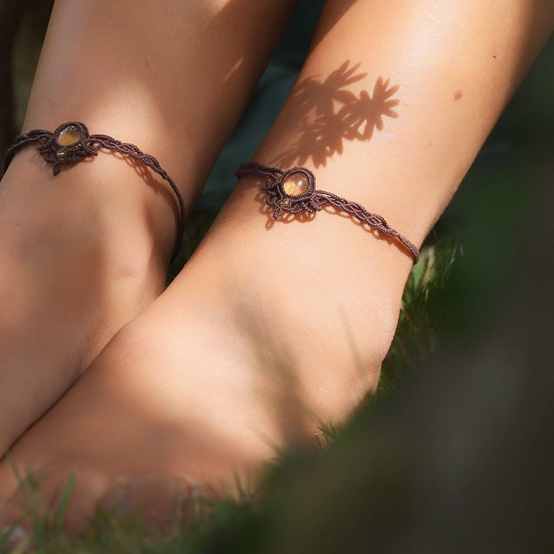 Bijoux de pieds, paire assortie de bracelets de chevilles à ambiance bohème celtique elfique. Fil marron foncé fin tissé en micro macramé avec entrelacs et arabesques, pierre centrale ovale petite taille jaune miel translucide. Bijoux hippie, bohème avec inspiration nature. Chevillères portées sur des chevilles à la peau pâle, bijoux de face avec jeu d'ombre et lumière et fleurs jaune en haut à droite.